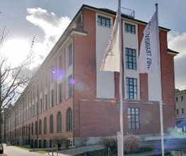 Außenansicht vom Schulungsgebäude an einem hellen Tag. Die Fassade ist rot und weiß geklinkert. Im Vordergrund sind 2 Fahnenmäste mit der Aufschrift „WBS TRAINING“ aufgestellt.
