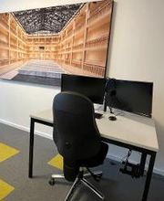 :Schreibtisch im Trainingsraum mit zwei Bildschirmen, einem Bürostuhl, Tastatur und Maus. An der Wand ein großes Bild eines Gebäudes von innen. 