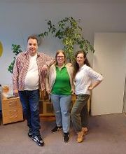 Ein Mann und zwei Frauen stehen nebeneinander und posieren für ein Foto. Neben ihnen befindet sich ein Schreibtisch mit Computerarbeitsplatz.