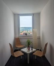 Ein kleiner Raum mit 3 Stühlen, Beistelltisch, Stehlampe und Fenster. Auf dem Tisch befindet sich eine kleine Topfpflanze.