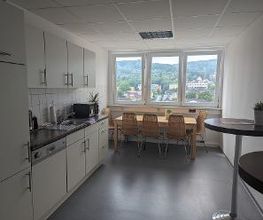 Eine weiße Küchenzeile mit Spülmaschine, Waschbecken und Mikrowelle. Hellbrauner Holztisch mit Stühlen steht vor einer Fensterfront.