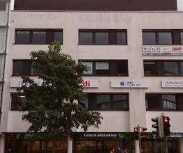 Ein mehrstöckiges weißes Gebäude mit dunklen Fenstern. An der Fassade sind Schilder von „ver.di“ und „WBS Training“ angebracht. Im Erdgeschoss befindet sich eine Bäckerei mit dem Schild „Frische Backwaren“. Vor dem Gebäude steht ein großer Baum.