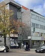 ußenansicht vom Gebäude an einer Straße mit Autos. Im Erdgeschoß gibt es einige Ladenlokale sowie eine Apotheke.