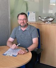 Ein Mann mit Brille und grauem Poloshirt sitzt an einem runden Tisch. Er hält einen Stift in der Hand und hat einen Klemmbrett-Block vor sich. Im Hintergrund steht ein Holzschrank, darauf eine silberne Fischfigur und ein eingerahmtes Zertifikat.