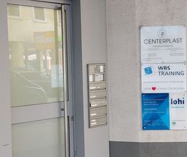 Eine Milchglastür zur Hausnummer 36. Rechts daneben befinden sich Briefkästen, Klingeln und einige Schilder von ansässigen Firmen, darunter WBS TRAINING.