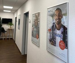 Flur mit mehreren Plakaten an der Wand. Links ein Tisch mit Pflanze und Barhocker. An der Wand darüber ein Bildschirm.