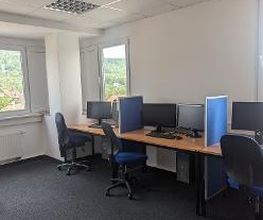 Schulungsraum mit mehreren Arbeitsbereichen, die durch blaue Trennwände voneinander abgegrenzt sind und mit PC-Bildschirmen, Tastatur, Maus und Headset ausgestattet sind. Blaue Bürostühle.