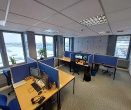 Raum mit mehreren Arbeitsplätzen mit jeweils 2 Bildschirmen, einem Bürostuhl, Tastatur und Maus. Im Hintergrund sind mehrere Schränke zu sehen, links eine Fensterfront mit Zimmerpflanzen. Der Boden ist mit Teppich ausgelegt.
