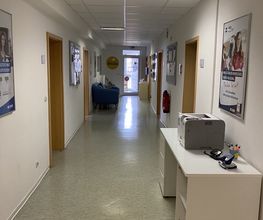 Flur mit mehreren Türen und mehreren Plakaten. Vorne rechts ein Regal mit Drucker, hinten links ein blaues Sofa.