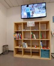 Holzregal mit Büchern. Darüber hängt ein TV, rechts steht ein Flyerständer, links ein Mülleimer.