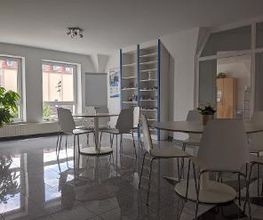 Aufenthaltsraum mit 2 Tischen und jeweils 5 Stühlen. Im Hintergrund sind 2 große Fenster und ein Bücherregal. Links steht eine Zimmerpflanze.