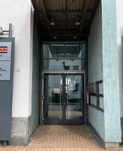 Das Bild zeigt den Eingang eines Gebäudes mit einer Glastür, darüber die schwarze Zahl „1“. Rechts und links sind graue Schilder: „Züblin“ (3. OG) und „WBS Training“ (2. OG). Der Boden ist gepflastert, die Wände hell, und es gibt eine Metallüberdachung.