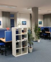 großer Raum mit mindestens 9 Arbeitsplätzen. Alle jeweils mit 2 Bildschirmen, einem Bürostuhl und Tastatur. Im Hintergrund sind mehrere Fenster, im Vordergrund stehen Regale. Mehrere Zimmerpflanzen sind im Raum verteilt. Blauer Teppichboden.