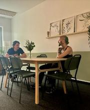 Pausenraum mit langem Tisch und 6 Stühlen. Im Hintergrund ein gelbes Sofa und an der Wand ein Regal mit 3 Bildern und einer Zimmerpflanze. 2 Personen sitzen am Tisch und unterhalten sich.