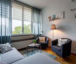 Aufenthaltsbereich mit 2 Sesseln und einem hellblauen Sofa. Links befindet sich eine Fensterfront mit blauen Vorhängen und einer Heizung. Rechts steht eine Pflanze. Mittig steht ein Beistelltisch mit Deko und eine Stehlampe.
