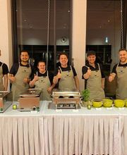 dean&david Catering | Stuttgart - Freundliches Catering Team hinter einer Buffetstation mit warmen Speisen, Saucen und frischen Kräutern für eine professionelle Eventbewirtung.