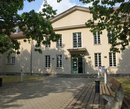 Außenansicht vom Gebäude. Helle Fassade mit weißen Fensterrahmen. Vor dem Gebäude ist eine Grünfläche mit Bäumen und einer Bank.