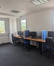 Schulungsraum mit mehreren Arbeitsbereichen, die durch blaue Trennwände voneinander abgegrenzt sind und mit PC-Bildschirmen, Tastatur, Maus und Headset ausgestattet sind. Blaue Bürostühle.