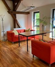 Pausenbereich an großen Fenstern mit zwei roten Sofas und Kissen und einem Tisch in der Mitte. Garderobenständer im Hintergrund. 