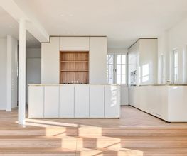 Offene Küche mit zurückhaltender Eleganz: Weiß, Holz und Messing prägen die reduzierte Gestaltung. Die großzügige Raumhöhe und das Tageslicht verstärken die architektonische Klarheit.