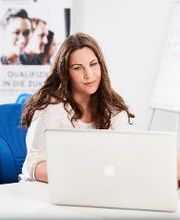 Dame, weiß gekleidet, dunkle lange Haare, sitzt mit einem dunkelhaarigen, weiß gekleideten Herrn am weißen Schreibtisch (blauer Schreibtischstuhl). Beide schauen gemeinsam in ein Notebook.