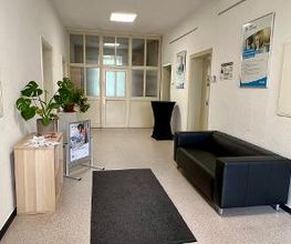ein Eingangsbereich mit einer schwarzen Couch auf der rechten Seite. Links steht ein Regal mit Flyern und einer Zimmerpflanze. Am Ende des Raums steht ein Stehtisch neben einer Glastür. Auf dem Boden liegt ein Teppich. 