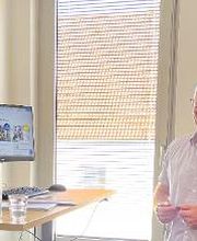 Ein Mann mit Brille und hellem Hemd steht lächelnd vor einem Stehpult. Auf dem Pult sind ein Monitor, eine Glaskaraffe, ein Glas und Unterlagen. Im Hintergrund sind Fenster mit Jalousien und das Dach eines Gebäudes zu sehen. Links steht eine Zimmerpflanze