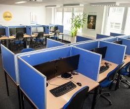 Ein Schulungsraum mit mehreren Arbeitsbereichen, die durch blaue Trennwände voneinander getrennt sind. Jeder Arbeitsplatz besteht aus 2 PC-Bildschirmen, Tastatur und Maus sowie Headset. Blaue Trennwände.