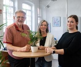 1 Mann und 2 Frauen stehen an einem Tisch mit Pflanze drauf. Alle 3 schauen freundlich in die Kamera. Neben ihnen steht eine weitere Pflanze.