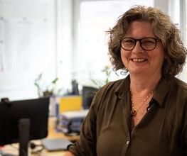 Eine Frau mit lockigem Haar, Brille und dunkelgrüner Bluse lächelt in die Kamera. Im Hintergrund sind ein Schreibtisch mit Monitor, Pflanzen und verschwommen Fenster mit Blick auf ein Gebäude zu sehen. Der Raum ist hell und freundlich eingerichtet.