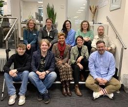 Gruppenbild vom Standortteam. 10 Personen sitzen im Flur auf einer Treppe und lächeln freundlich in die Kamera.
