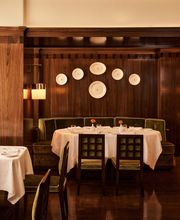 A large round dining table set with white linen and green velvet chairs under a dark wood wall adorned with a gallery arrangement of ceramic plates.