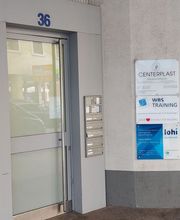 Eine Milchglastür zur Hausnummer 36. Rechts daneben befinden sich Briefkästen, Klingeln und einige Schilder von ansässigen Firmen, darunter WBS TRAINING.