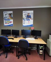 Arbeitsplätze mit jeweils einem Bürostuhl, 2 Bildschirmen und Tastatur und Maus. Links eine Zimmerpflanze, rechts ein Regal mit Drucker. An der Wand 2 Plakate der WBS TRAINING.