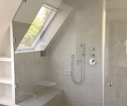 Dusche unter einer Dachschräge mit großem Fenster für natürliches Licht und minimalistischen Armaturen. Die Wände, die Sitzbank in der Dusche und die Einbaufächer sind hier ebenfalls fugenlos gestaltet.