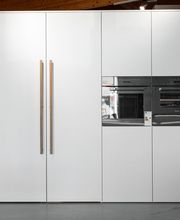 Elegante, moderne Hochschrankküche mit großflächigen weißen Fronten, integrierten schwarzen Einbaugeräten und einer schlanken Glasfront. Die grifflosen Schrankelemente mit langen Holzgriffen verbinden minimalistisches Küchendesign mit warmen natürlichen Akzenten. Die sichtbare Holzbalkendecke ergänzt die hochwertige, zeitlose Küchengestaltung.