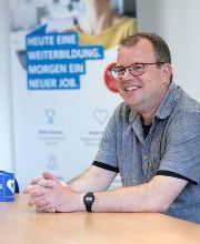 Ein Mann mit Brille und grauem Hemd sitzt an einem Tisch. Vor ihm steht eine blaue Kaffeetasse.