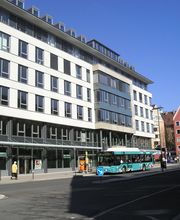 Ein vierstöckiges, weißes Gebäude an einer Straße. Vor dem Gebäude hält gerade ein Linienbus.