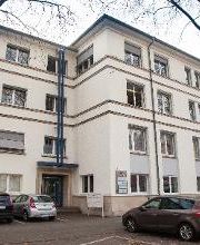 Außenansicht auf das Gebäude von der gegenüberliegenden Straßenseite. Hier wird der Eingang präsentiert. Es gibt einige Parkplätze vor dem Gebäude.