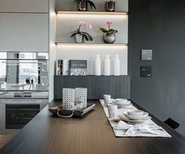 Moderne Designküche mit großzügiger Kochinsel und integrierter Sitzgelegenheit. Die Kombination aus matten Fronten, dunkler Holzoptik, offenen Regalen mit indirekter LED-Beleuchtung und stilvoll gedecktem Esstisch schafft eine elegante und wohnliche Atmosphäre. Hochwertige Einbaugeräte sowie durchdachte Stauraumlösungen machen diese LEICHT-Küche zu einem perfekten Beispiel für modernes Küchendesign.
