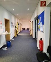 Ein heller Flur mit einer blauen Pinnwand an der Wand, sowie Türen, einem Feuerlöscher und Sitzgelegenheiten.