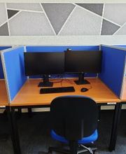 Arbeitsplatz bestehend aus 2 PC-Bildschirmen, Tastatur, Maus und Headset sowie einem blauen Bürostuhl und blauen Trennwänden.