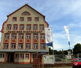 Ein Bild der Außenansicht des Kooperationsstandortes in Grimma. Das Haus hat eine beige Fassade und rote Ränder. Es gibt einen roten Schriftzug „Haus der Wirtschaft“.