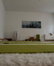 Der Yogaraum von Saccidananda-Yoga vom Altar aus. Unter dem Bild mit den Räucherstäbchen liegen Blöcke, Gurte und noch 2 Yogamatten. Der helle und freundliche Raum fasst regulär 16, maximal 20 Yogis.