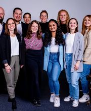 Gruppenbild vom Team. Die Personen lächeln alle freundlich in die Kamera. Ein Mitarbeiter hält einen Hund auf dem Arm.