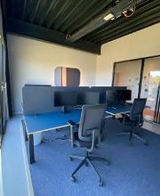 Schreibtische im Trainingsraum mit zwei Bildschirmen, Tastatur, Maus und Headset. An der Wand hängen Akustikpaneele, große Fensterfront in die Natur sowie in den Flurbereich