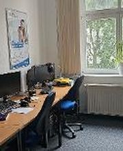 Schulungsraum mit 5 Arbeitsplätzen mit jeweils 2 Bildschirmen, einem Bürostuhl, Tastatur, Maus und Headset. An der Wand ein Kalender und ein Plakat. Auf dem Fensterbrett eine Pflanze.