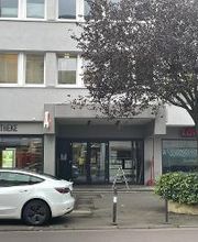 Eingangsbereich eines Bürogebäudes, dvor eine Straße und ein geparktes Auto. Links im Erdgeschoss befindet sich eine Apotheke.