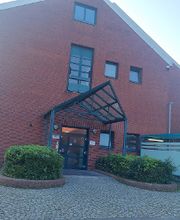 Außenansicht vom Gebäude. Backsteingebäude mit einer dunkelgrünen Eingangstür und Überdachung. Links sind mehrere Unternehmensschilder befestigt, unter anderem auch eines von WBS SCHULEN.