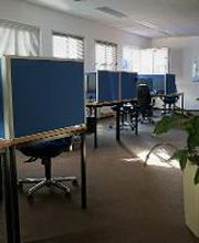 Schulungsraum mit mehreren Arbeitsbereichen, die durch blaue Trennwände voneinander abgegrenzt sind und mit PC-Bildschirmen, Tastatur, Maus und Headset ausgestattet sind. Fensterreihe und Bilder an der Wand.